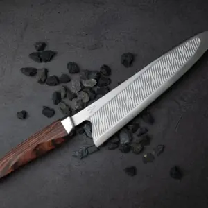 Ein RYVON Knives Kochmesser mit gemusterter Klinge und Holzgriff liegt auf einer dunklen Fläche mit verstreuten schwarzen Steinen.